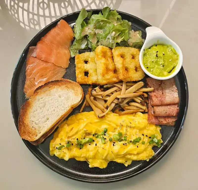 A breakfast plate with smoked salmon, scrambled eggs, hash browns, bacon, mushrooms, salad, avocado and toast