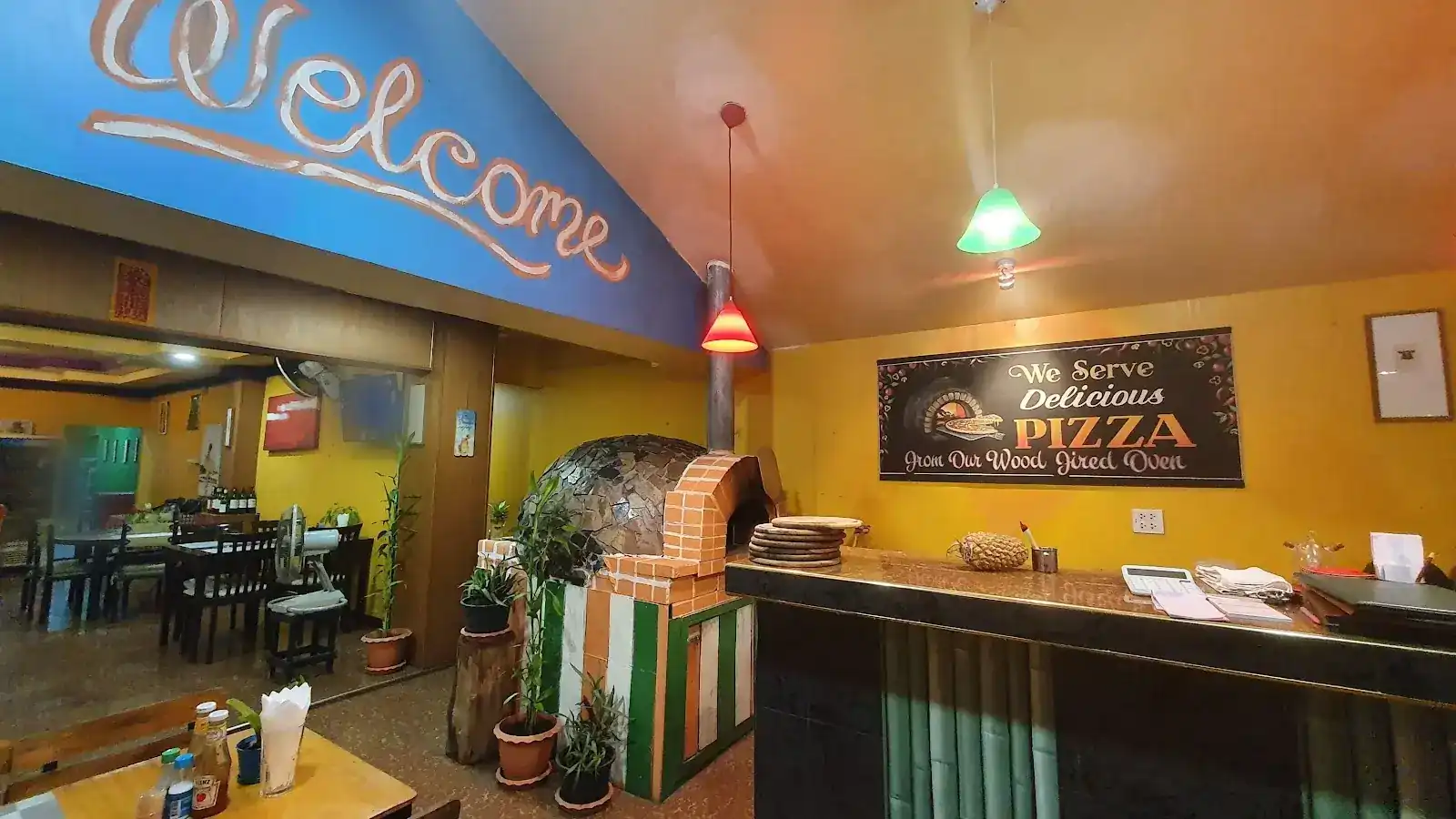 Inside the restaurant: Welcome painted by hand on the blue ceiling beam, the wood-fired oven with its stone dome, and the counter against the yellow wall