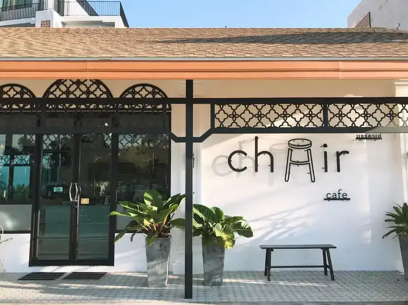 The white front of Chair Cafe with black iron lattice, potted plants, a bench and the chair sign on the wall