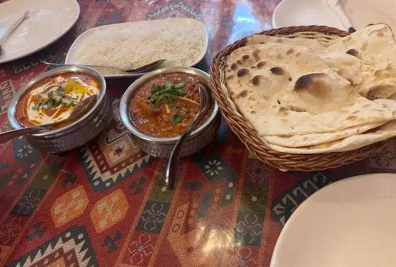 Two curries in hammered steel bowls, white rice and a basket of naan on a patterned tablecloth