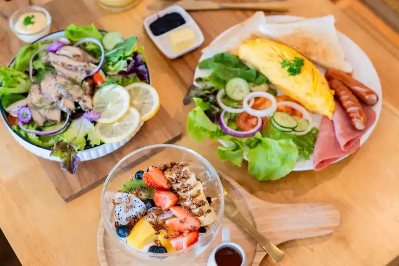 A grilled chicken salad, an omelette breakfast plate with sausages and toast, and a fruit yogurt bowl