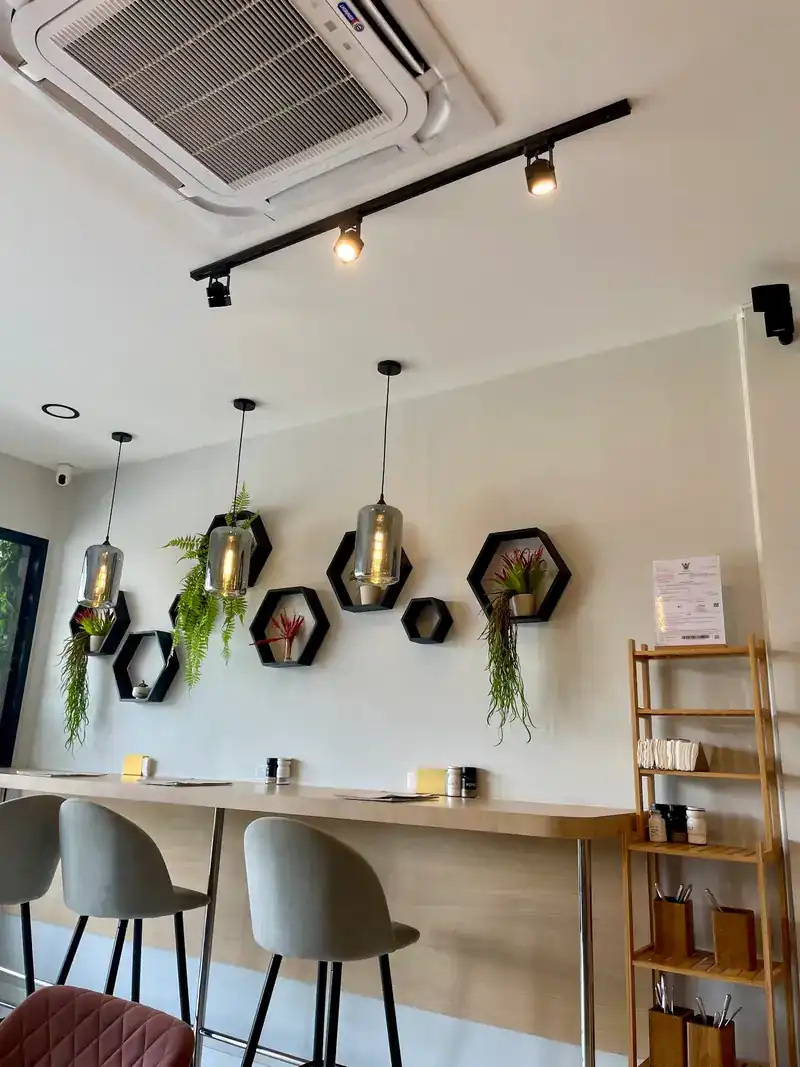 A long wooden counter with grey stools under hexagon shelves with hanging plants and glass lamps