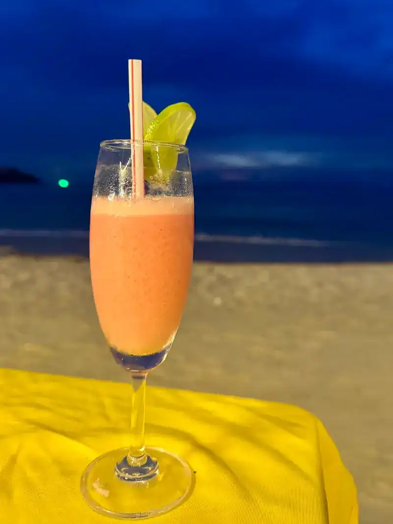 A pink blended drink with lime in a tall glass on a yellow table, the sea behind it at dusk.