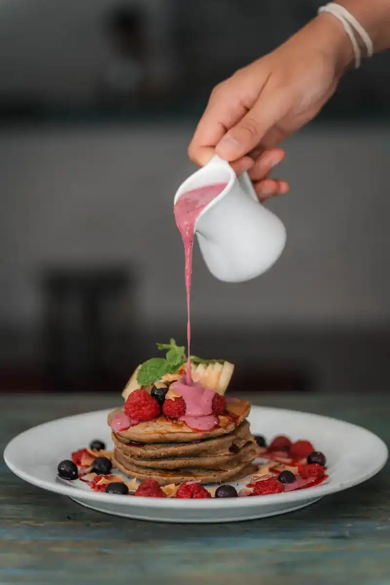 A hand pouring pink berry sauce over a stack of pancakes