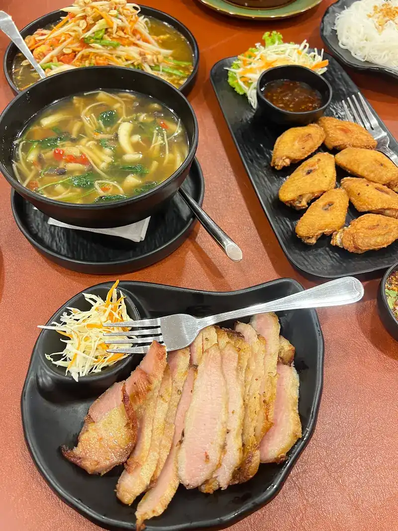 Papaya salad, a bowl of spicy soup, fried chicken wings and sliced grilled meat on one table
