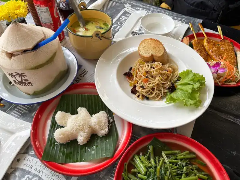 One table: a young coconut, a small enamel pot, noodles, satay, stir-fried morning glory and rice shaped like a bear