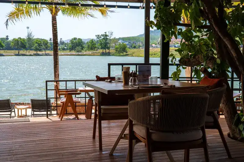 A table and woven chairs on the deck, with the lake and palms beyond