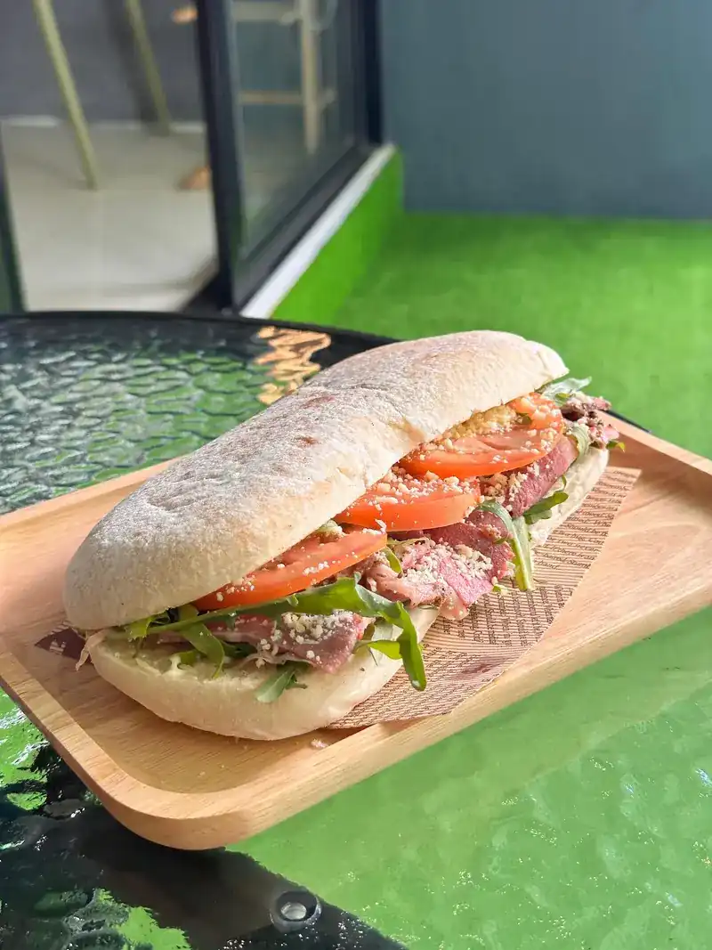 A ciabatta sandwich with sliced meat, tomato and greens on a wooden board