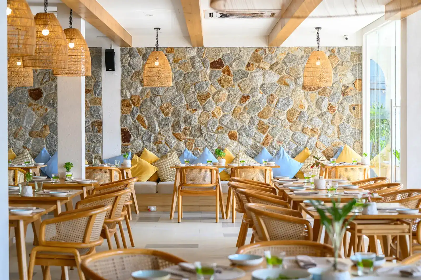 The dining room: rattan chairs and wooden tables under woven lamps, in front of a wall of rough stone
