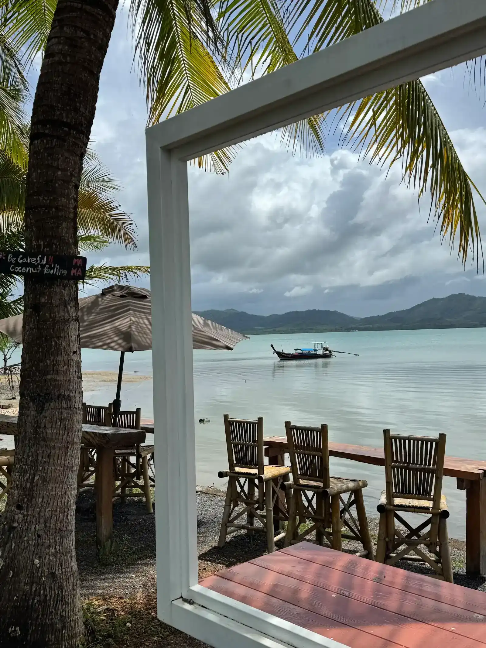 Looking through the white frame on Mama Kitchen's shore to a longtail boat on the bay, with bamboo chairs at the water's edge