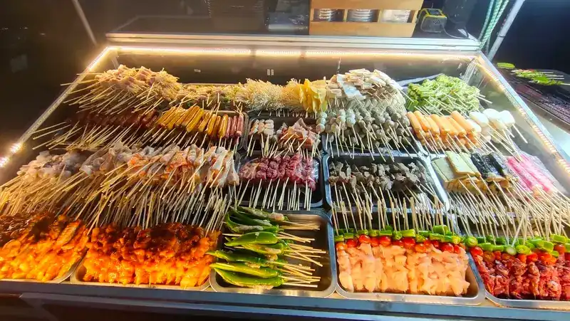 The lit display case at night, full of skewers of meat, seafood, sausages, mushrooms and vegetables