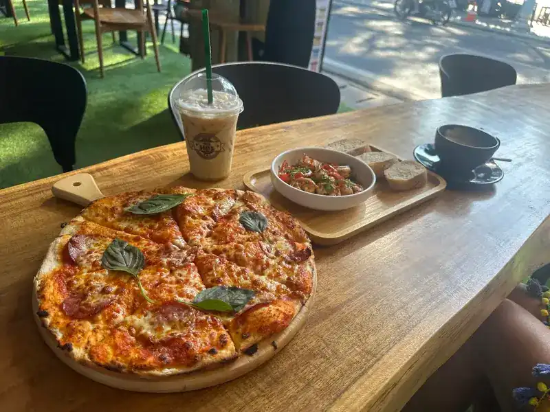 Thin pizza topped with sliced sausage and basil on a wooden counter, with an iced coffee and a cup of coffee