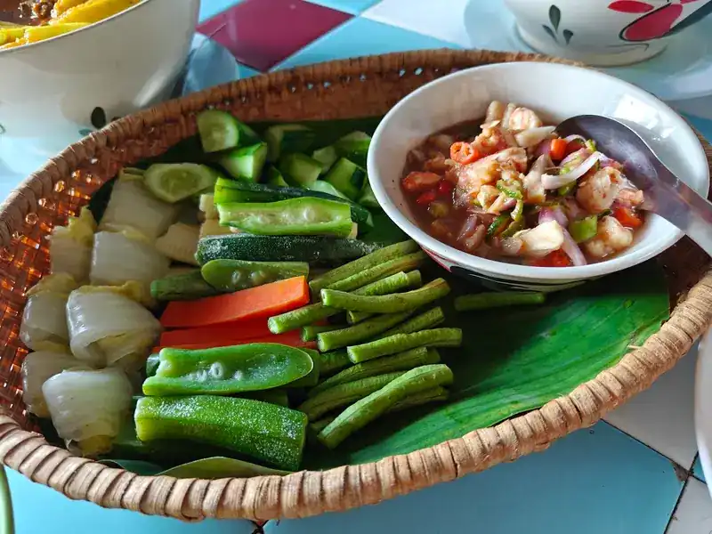 A woven basket of fresh vegetables with a bowl of prawn, tomato and chilli relish