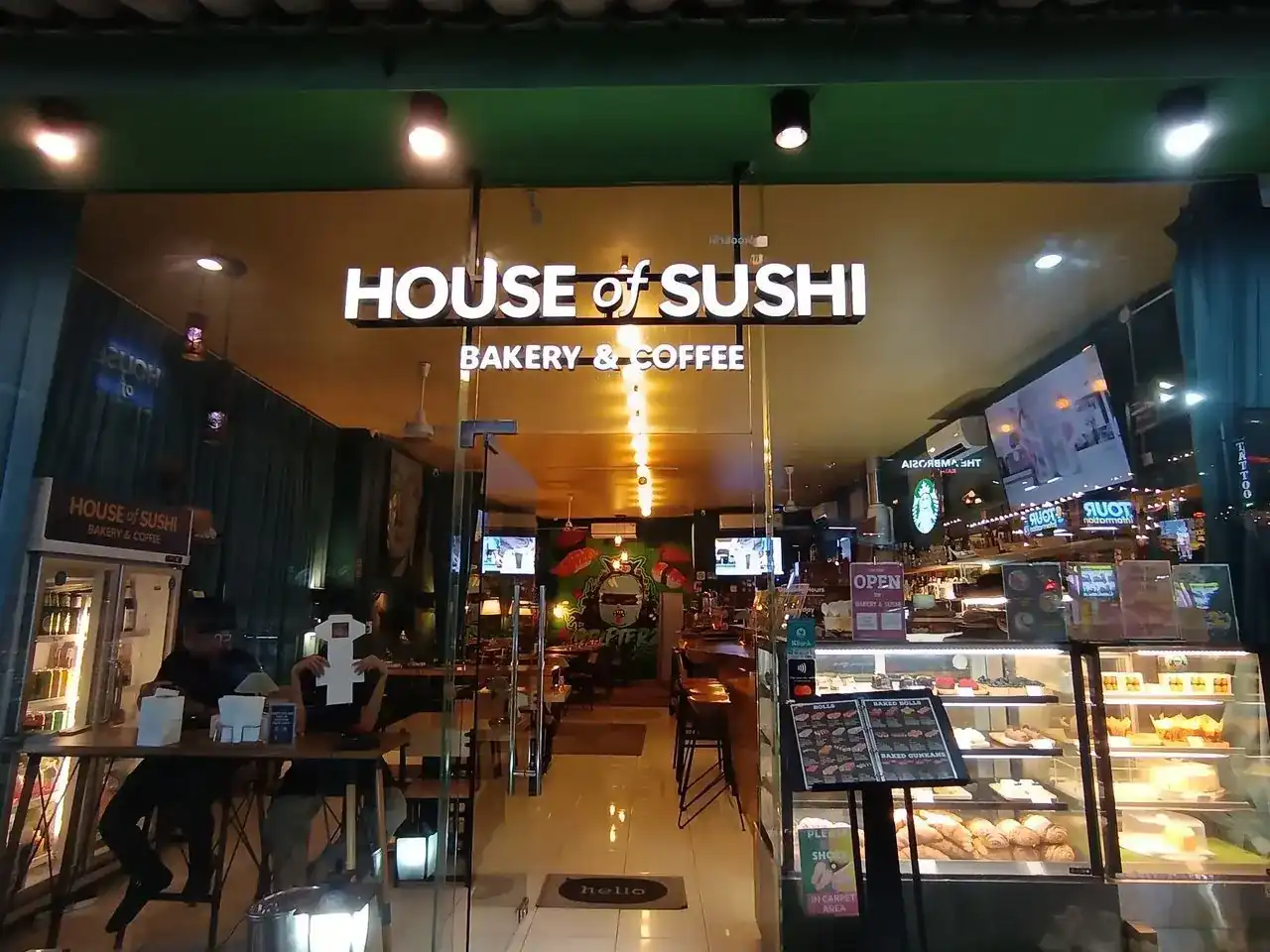 The shopfront after dark: the lit House of Sushi sign over a glass door, with a bakery display case beside it