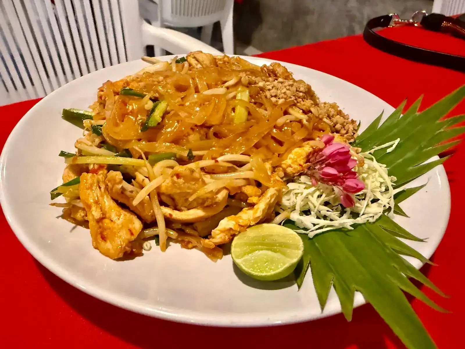 Pad thai on a white plate with lime, shredded cabbage, a fan of cut leaf and pink flowers, on a red tablecloth