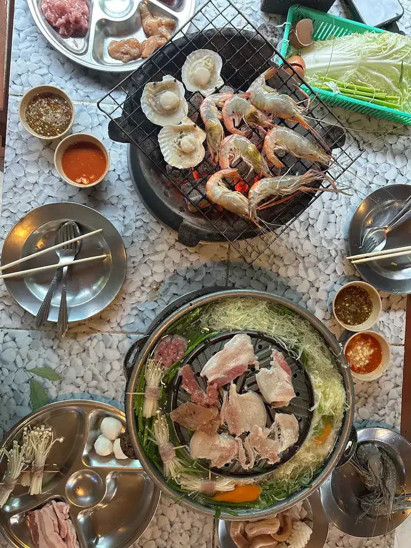 A table seen from above: scallops and prawns on a charcoal grill, sliced pork on a mookata pan, small bowls of dipping sauce