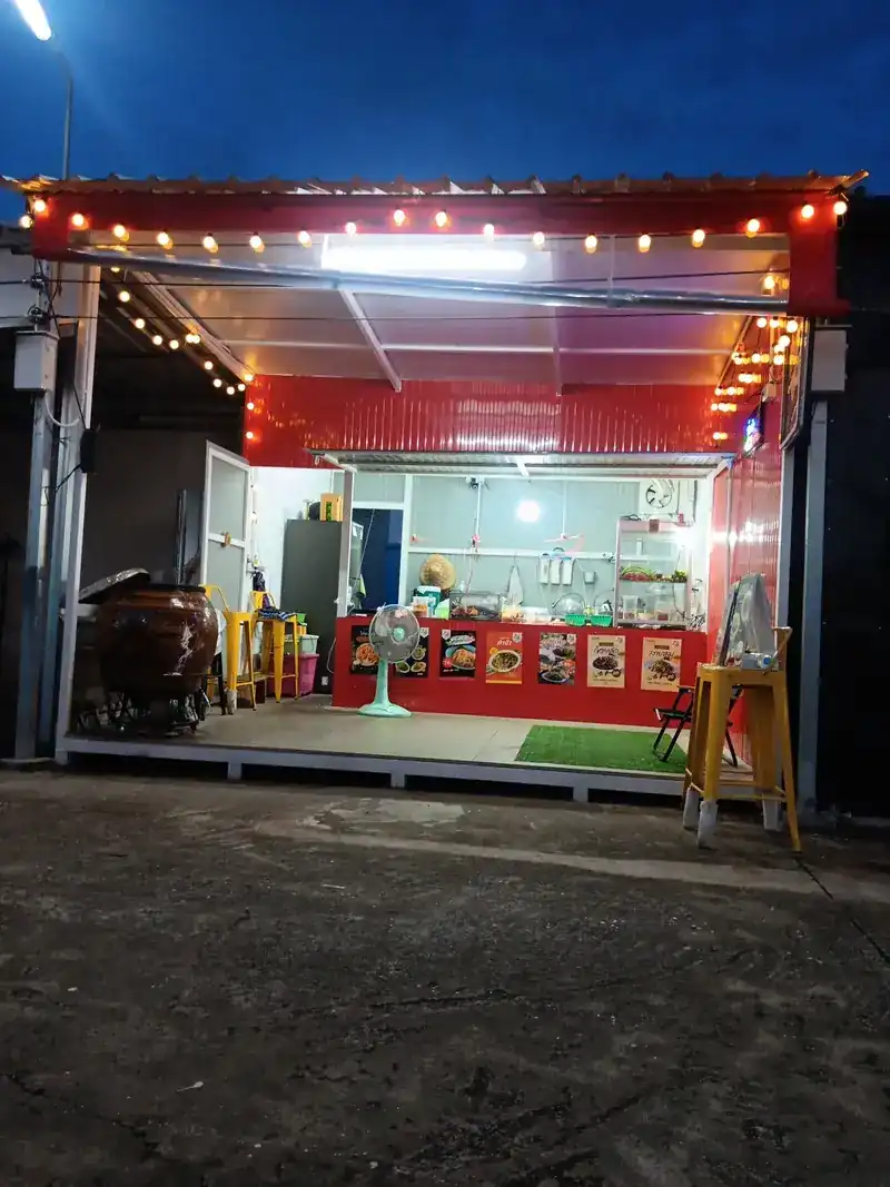 The stall after dark: red walls, string lights, yellow stools and a big glazed jar out front