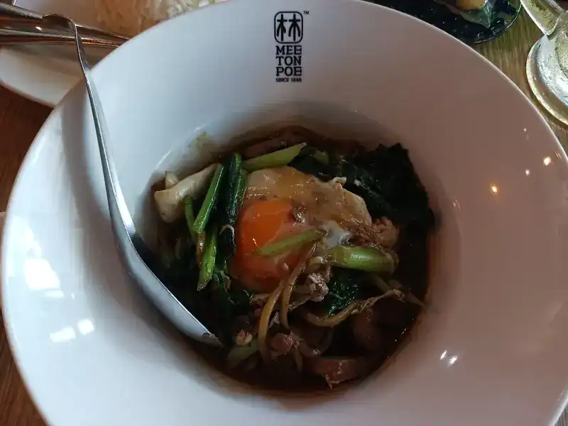 Noodles with greens, sliced meat and a fried egg in a Mee Ton Poe bowl