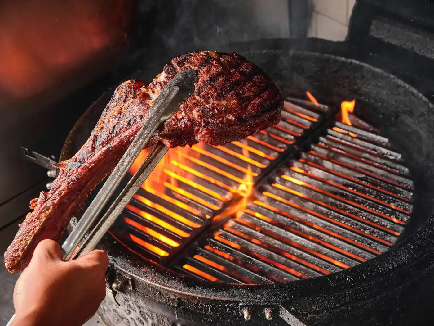 A long-bone steak lifted with tongs off a glowing charcoal grill