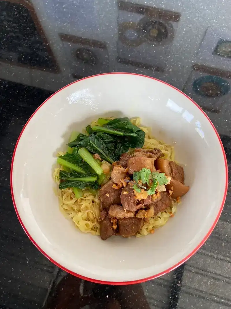 Braised pork on flat egg noodles with Chinese kale in a red-rimmed bowl
