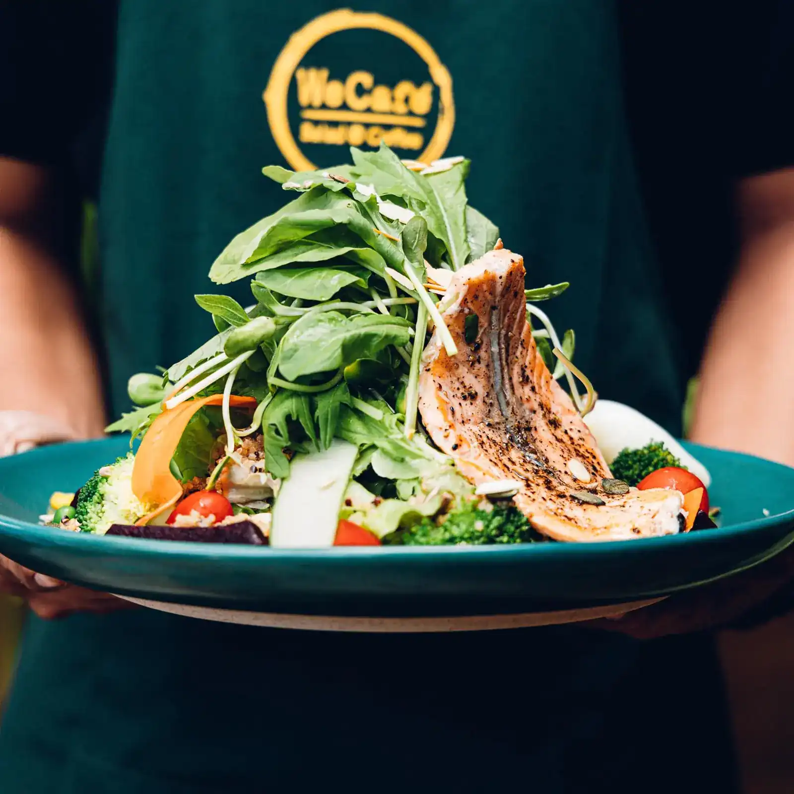 A salad piled high with greens and grilled salmon, held out on a teal plate in front of the green We Cafe' apron