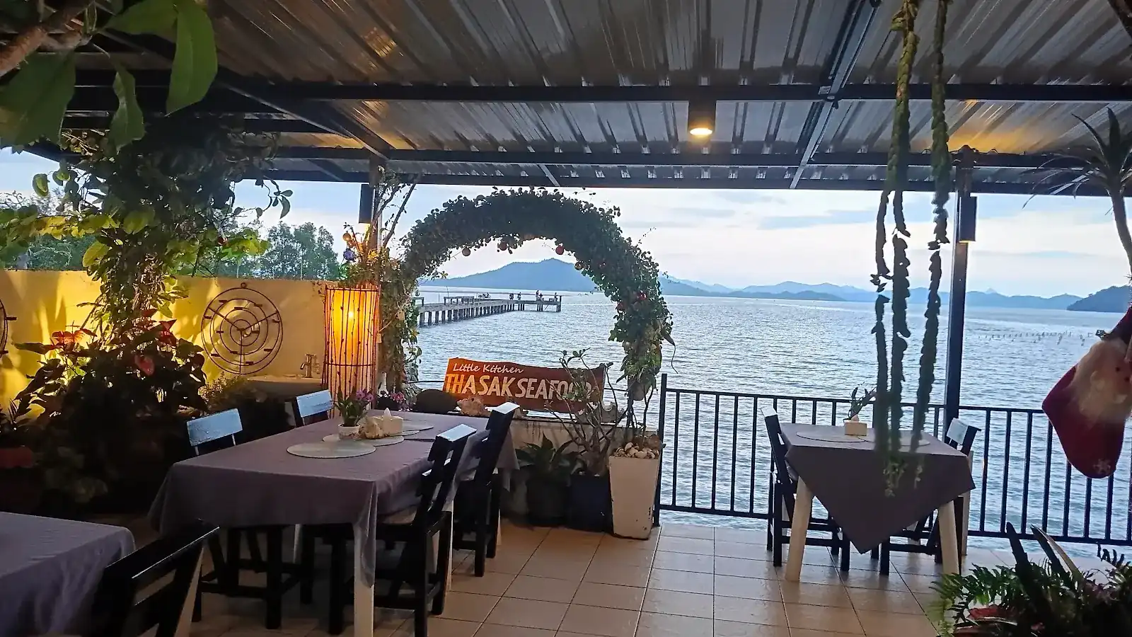 The deck at dusk: tables in lilac cloth, the Little Kitchen Thasak Seafood sign under a leafy arch, a pier and islands across the sea