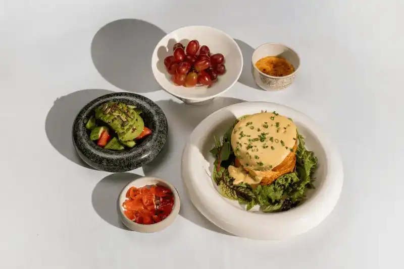 A breakfast spread over five dishes: a croissant under sauce on salad leaves, avocado, salmon, grapes and a small dessert
