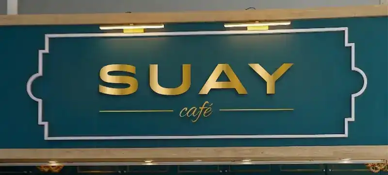 The teal SUAY café sign with gold letters above the counter / ป้ายร้าน SUAY café สีเขียวหัวเป็ดตัวอักษรสีทองเหนือเคาน์เตอร์