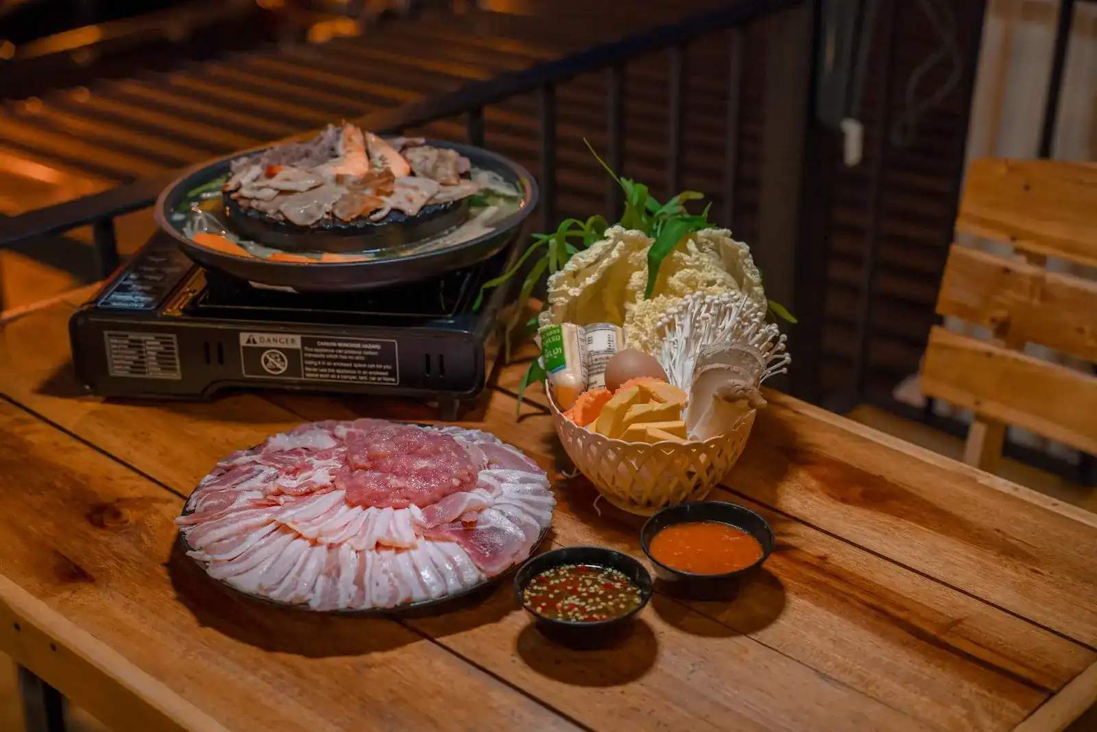 A mookata pan with pork and prawns on a gas burner, a platter of sliced pork, a basket of vegetables and two dipping sauces on a wooden table