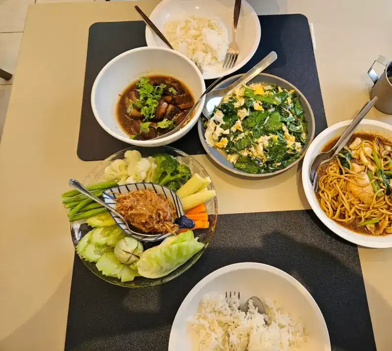A table of local dishes: braised pork, greens stir-fried with egg, a chilli dip with fresh vegetables, and yellow noodles