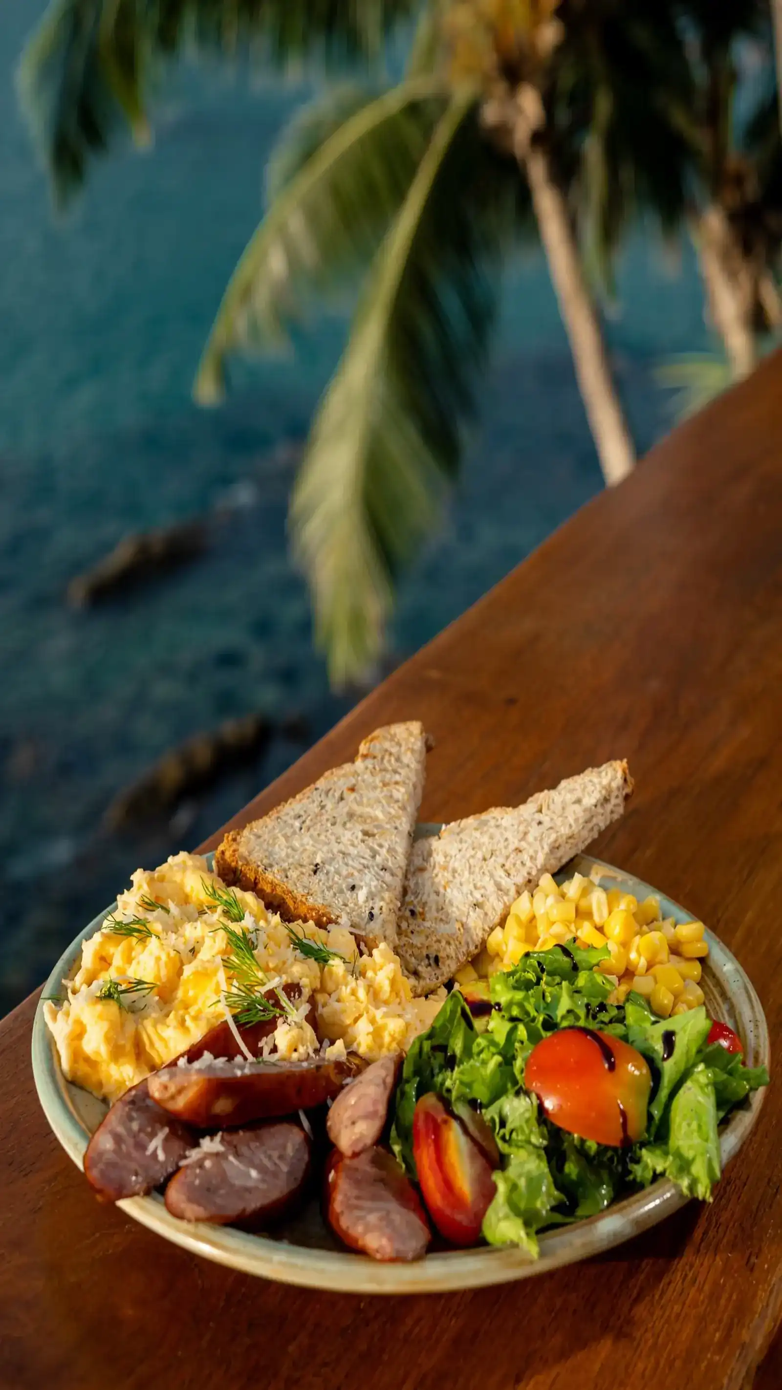 Scrambled eggs, grilled sausage, salad, sweet corn and toast on a wooden rail above the sea