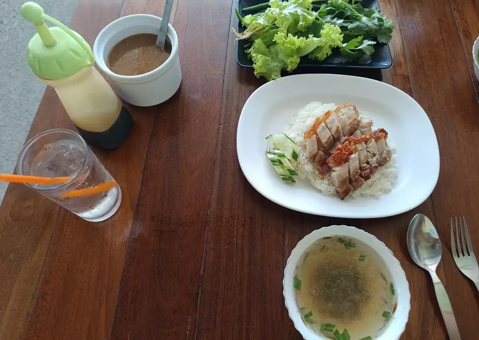 Crispy pork on rice with cucumber, a bowl of clear soup, fresh greens and dipping sauce on a wooden table