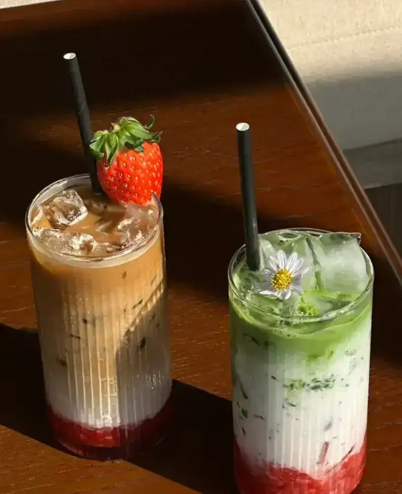 Two iced drinks on a sunny table, one topped with a strawberry, one green with a daisy
