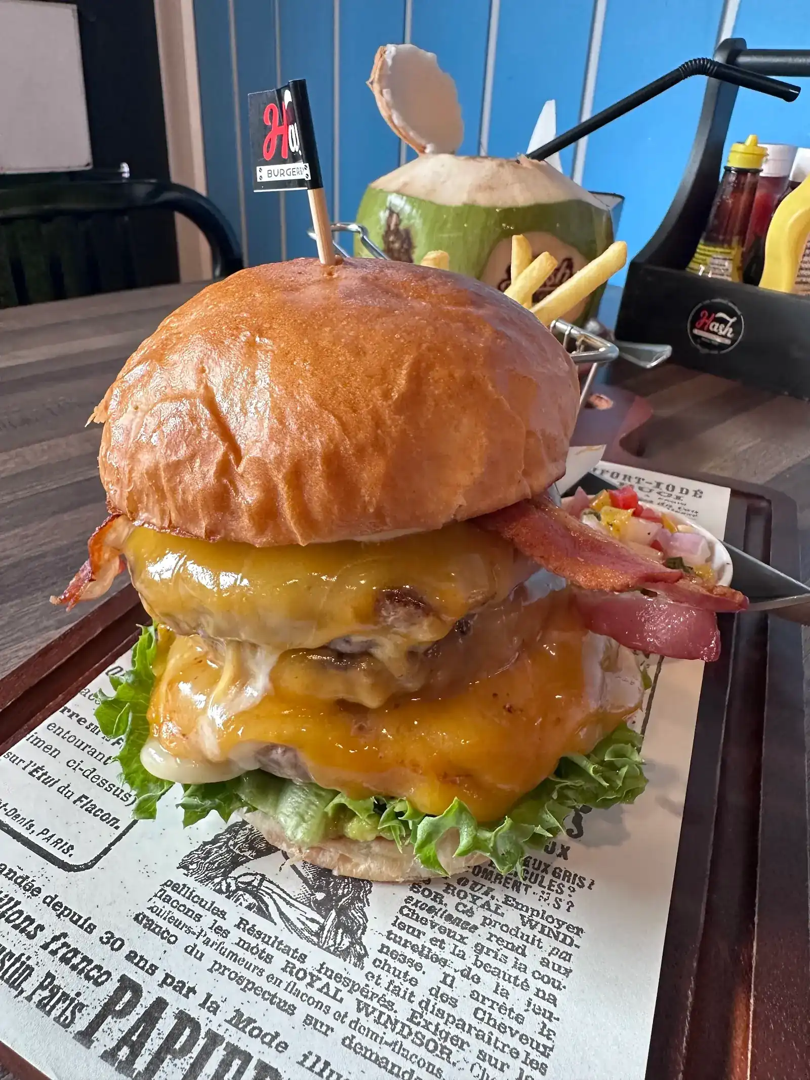 A double cheeseburger with bacon and lettuce in a toasted bun, on a board with fries and a fresh coconut behind