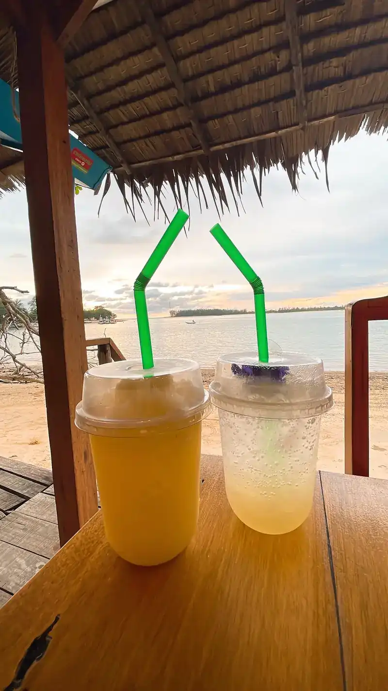 Two cold drinks on a wooden table under a thatched roof, with the beach behind