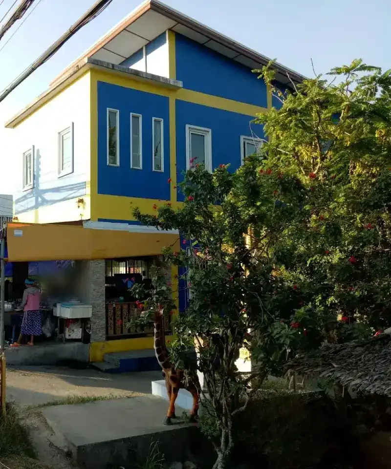 The blue-and-yellow shop house by the road, a red-flowered tree and a wooden giraffe in front