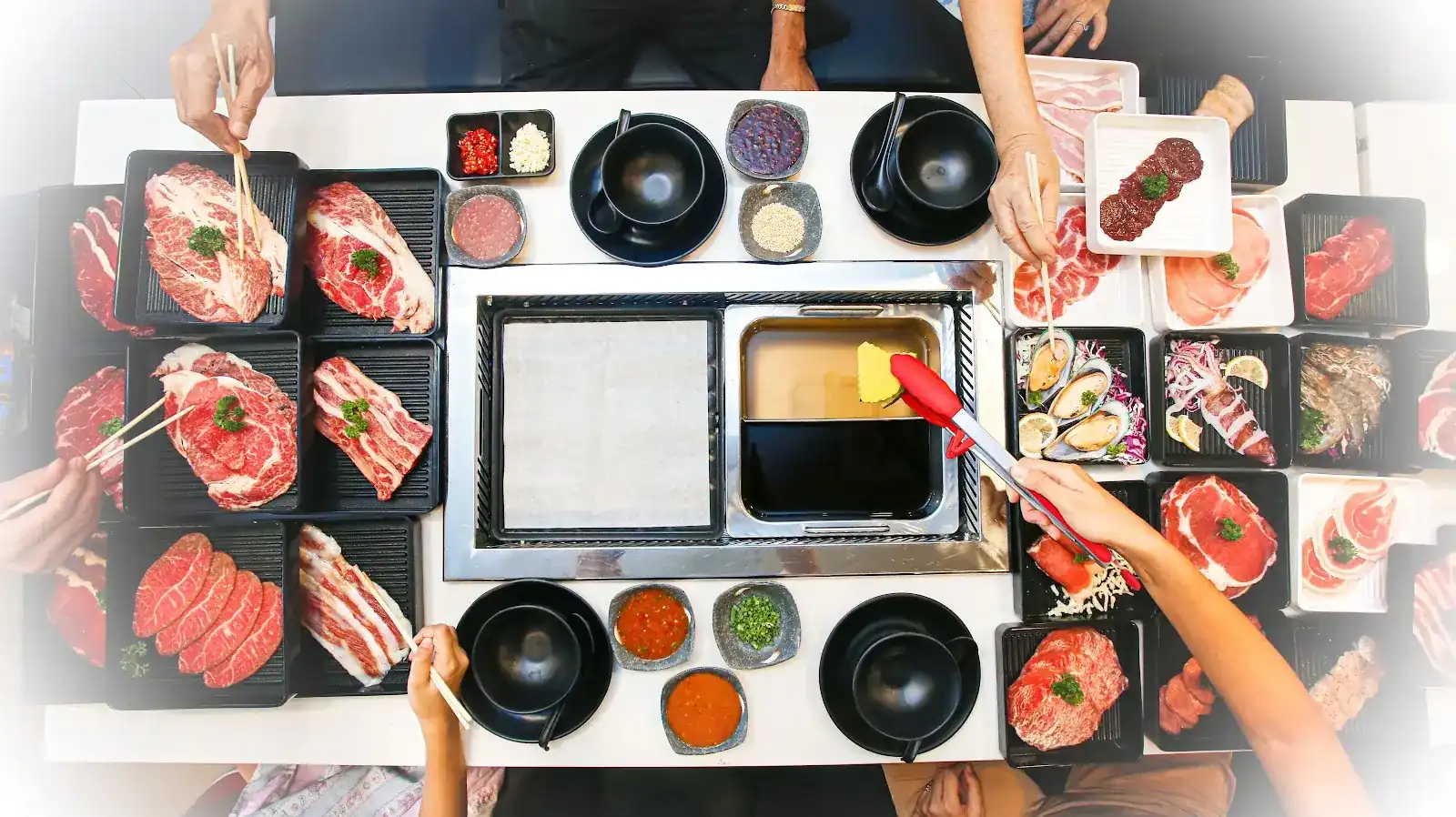 Overhead view of a table at Panda Shabu: marbled beef and pork on black trays around the built-in grill plate and shabu pot