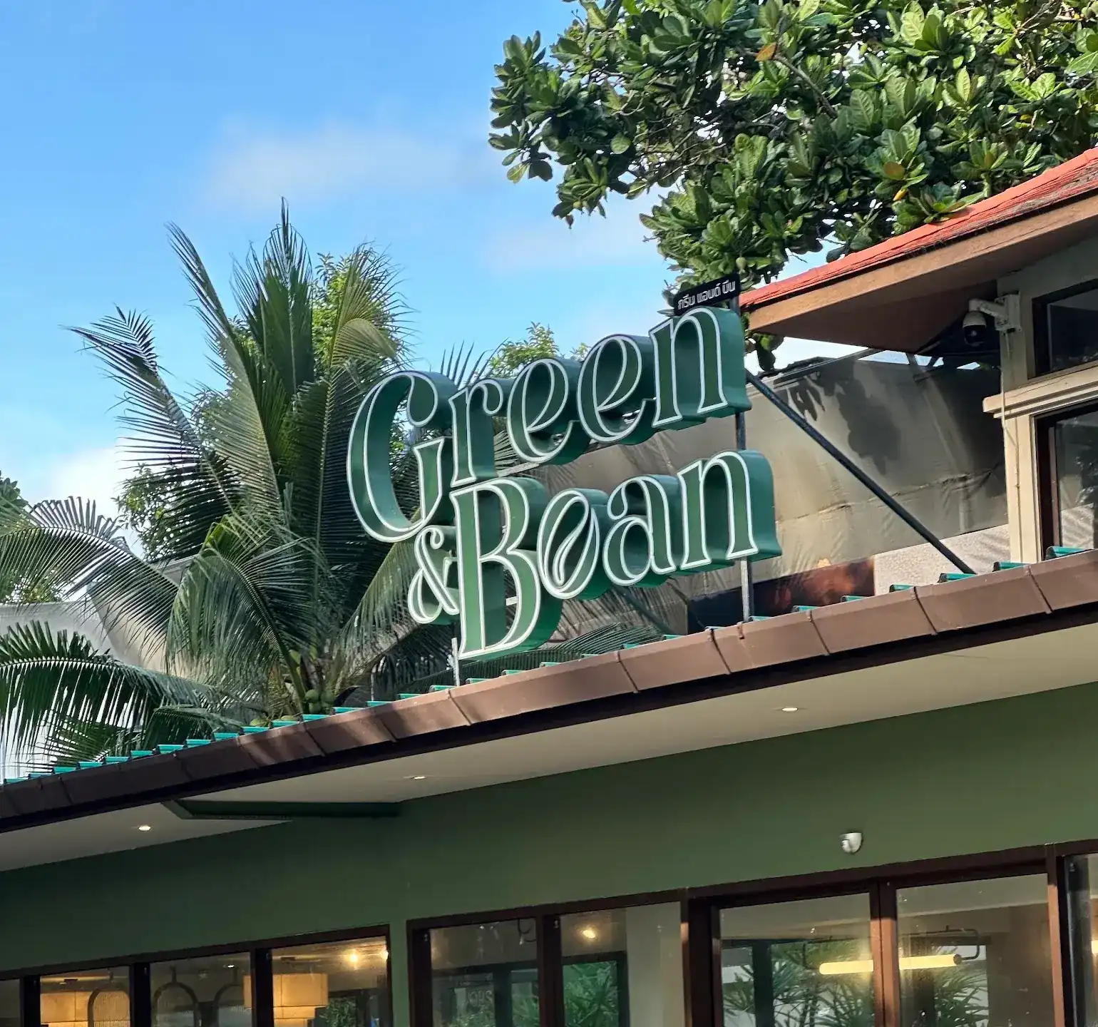 The green Green & Bean sign on the café roof, with palm trees and blue sky behind