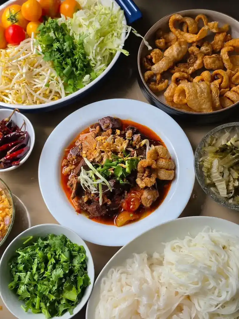 A plate of red nam ngiao broth with pork and crisp pork rind, surrounded by bowls of rice noodles, bean sprouts, cabbage, herbs, tomatoes and dried chillies