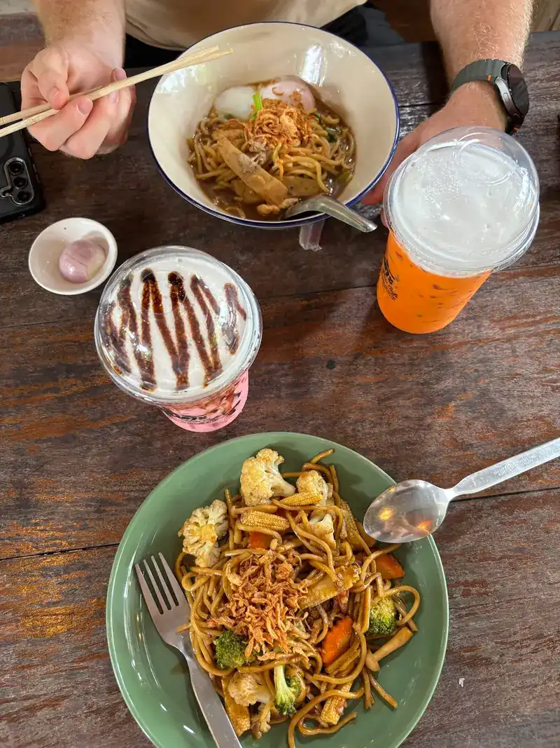 Seen from above: fried noodles with vegetables, a bowl of noodles and two iced drinks on a wooden table