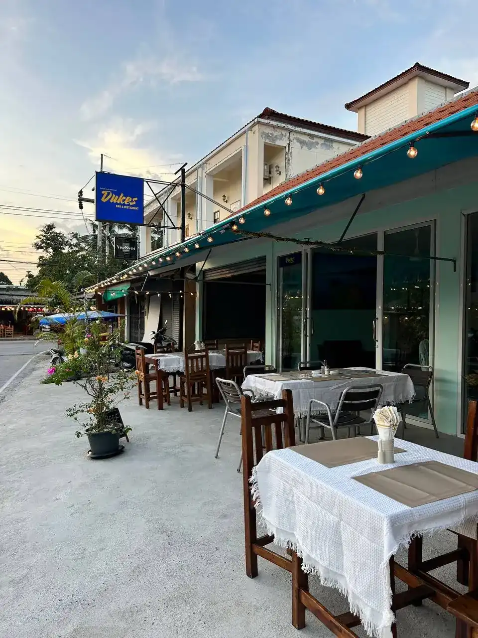 Duke's terrace at dusk: tables in white cloths under a teal awning with string lights, and the blue Dukes sign by the road