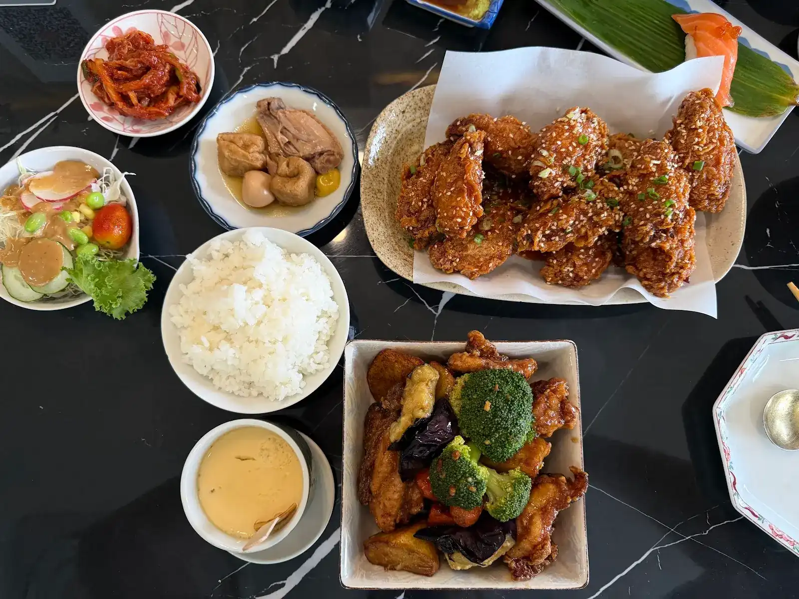 A table seen from above: glazed fried chicken, a stir-fry with broccoli and eggplant, rice, kimchi, salad, oden and steamed egg on black marble
