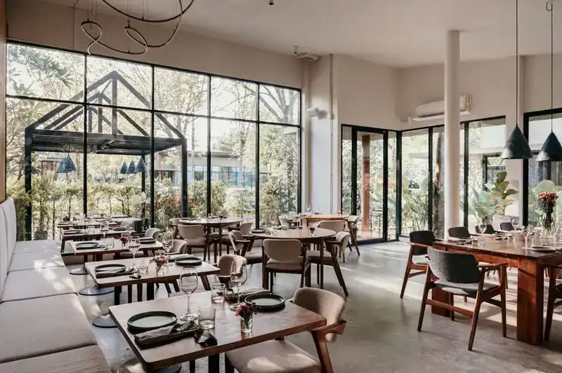 Bright dining room with wooden tables, tall windows onto a garden and a looping light sculpture overhead