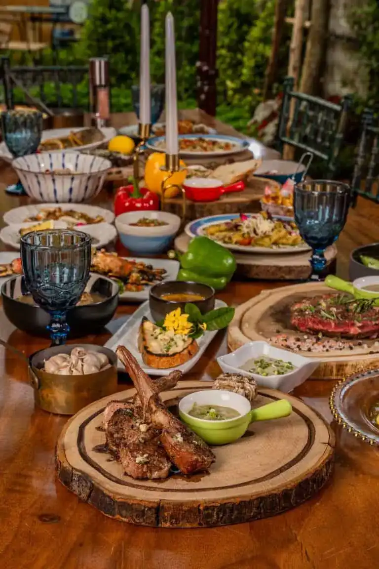 A wooden garden table covered with dishes: lamb chops on a wooden board, steak, bowls of sauce, blue goblets and candles