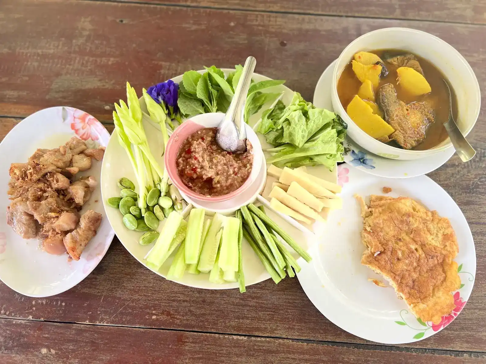 Nam prik with a platter of fresh vegetables, fried pork, an omelette and a fish curry with pumpkin on a wooden table