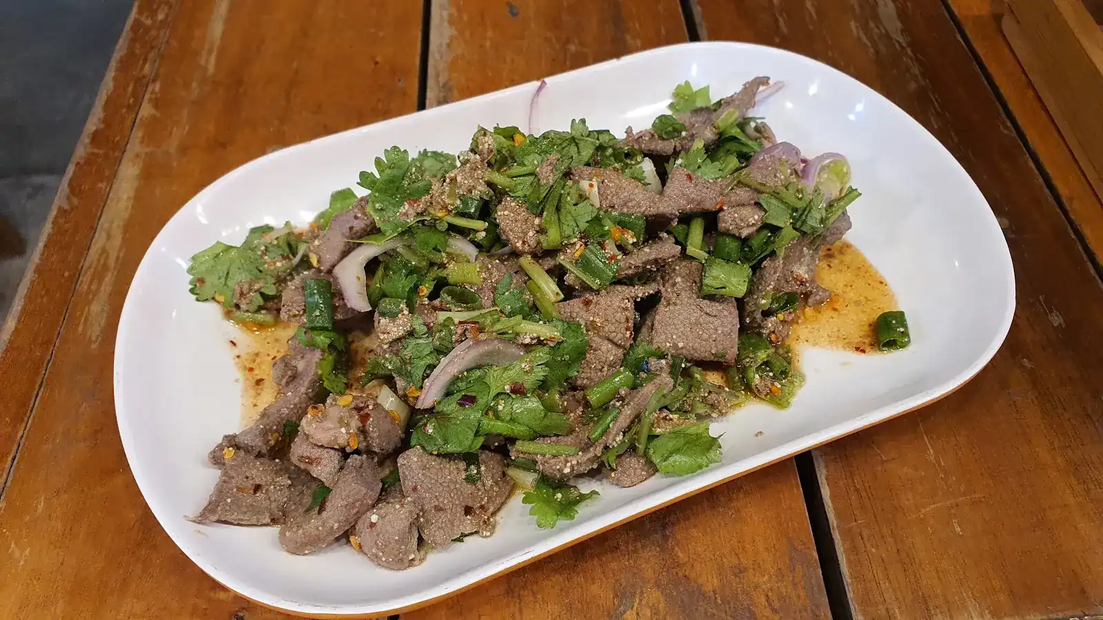 Larb of sliced meat with coriander, spring onion and red onion on a white plate, on a wooden table