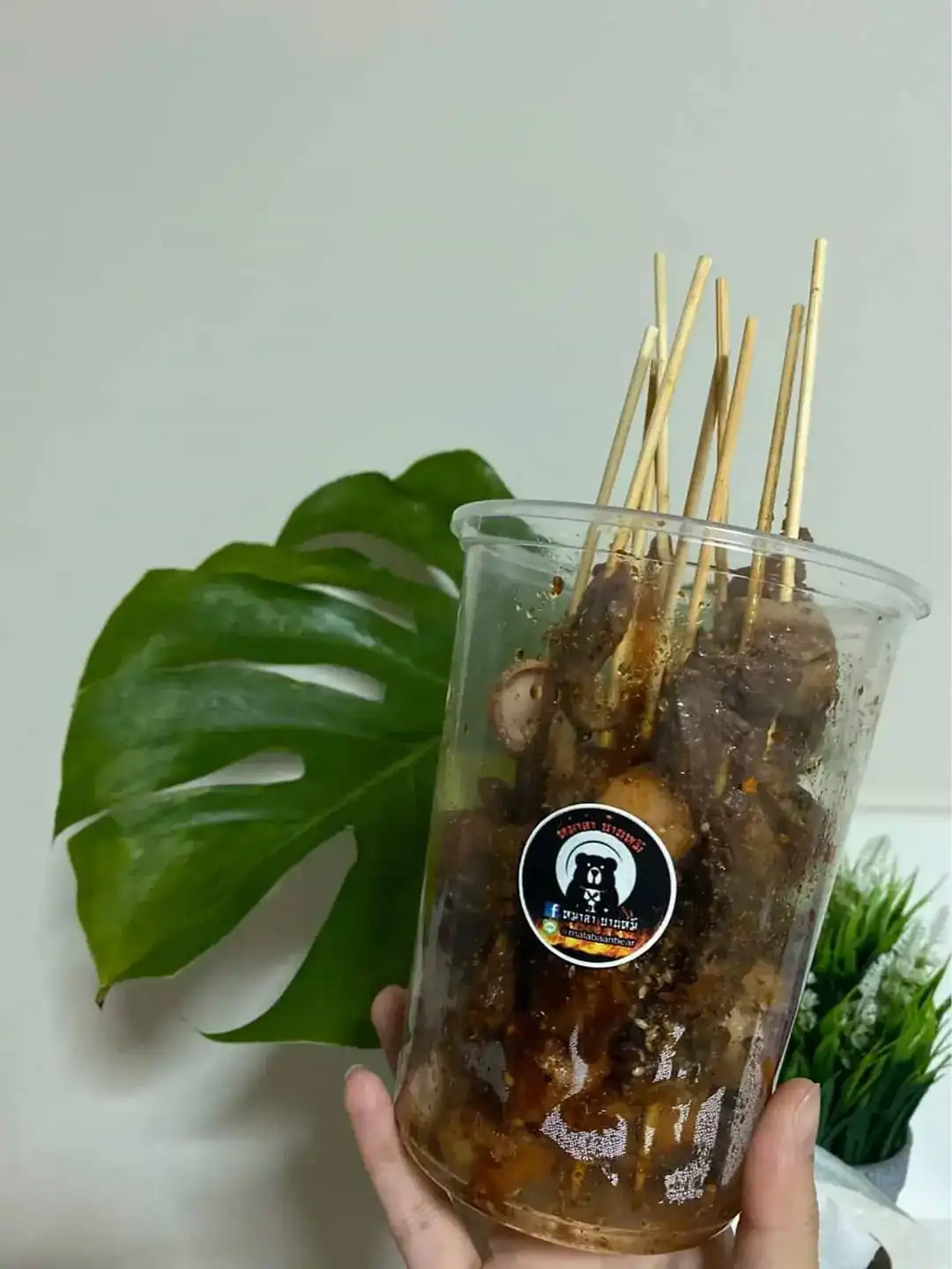Mala skewers packed in a tall clear cup, held up by hand, with the bear sticker of Mala Baan Mee
