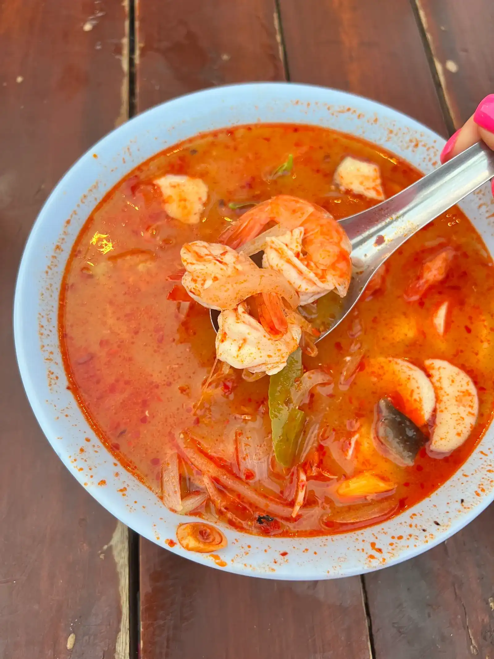 A bowl of tom yum with prawns, mushrooms and chilli, a spoon lifting the prawns, on a wooden table