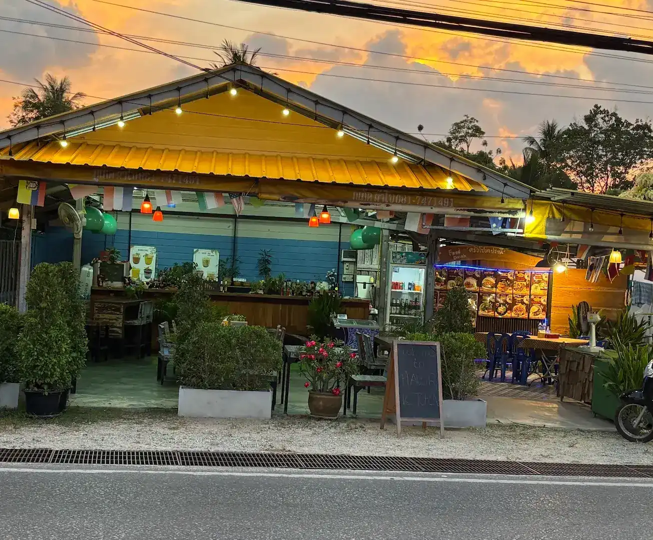 The front of Favorite Kitchen at sunset: a yellow tin roof, string lights and potted plants beside the road