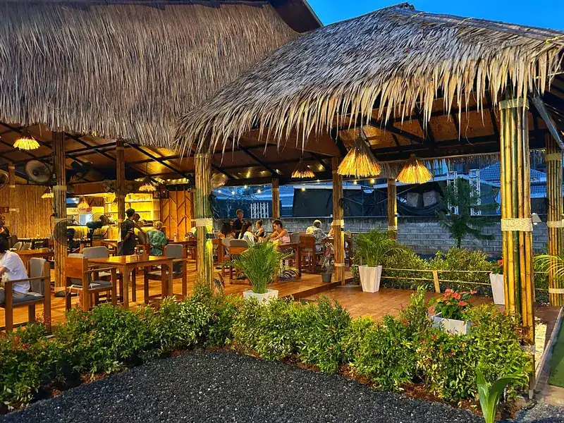 The bamboo and thatch dining pavilion at dusk, lamps lit, with a planted border in front